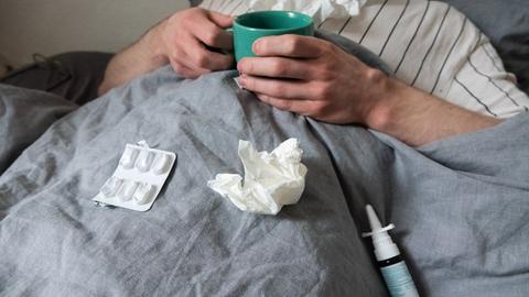 Ein erkälteter Mann im Bett, auf der Bettdecke liegen Taschentücher, Nasenspray und ein Blister mit Tabletten