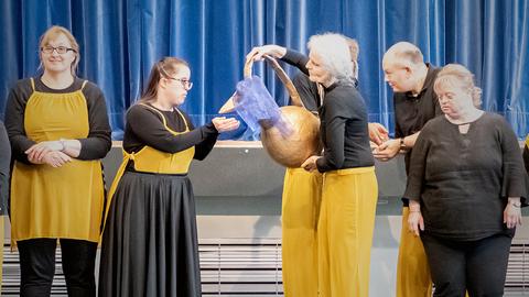 Bühnenszene: Eine Frau schenkt aus einer großen Kanne aus, eine andere hält die Hände geöffnet