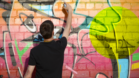 Ein Mann der vor einer Wand steht, eine Spraydose in der Hand hat und an diese Wand ein Grafitti sprüht. Man sieht den Mann von hinten